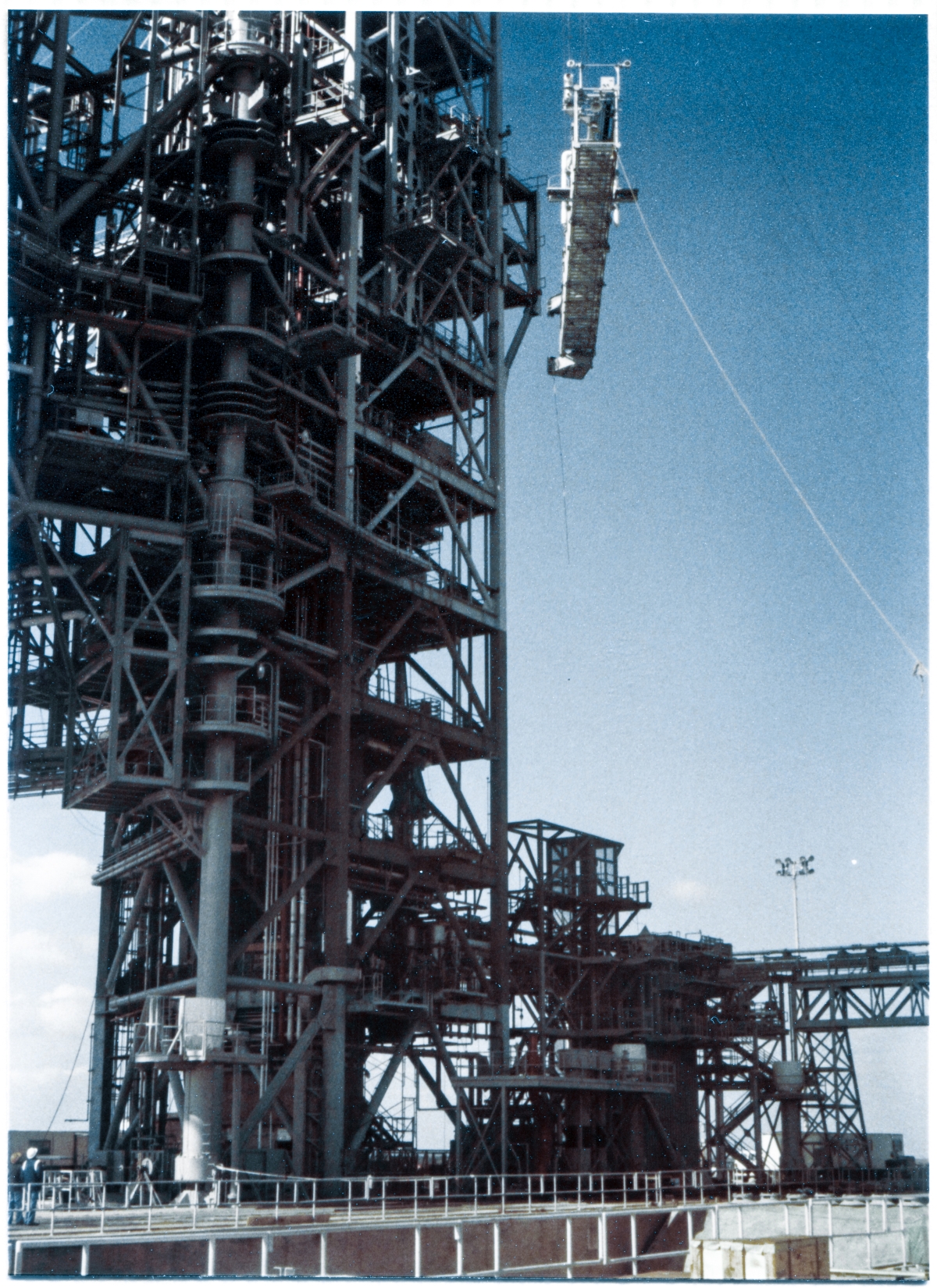 Image 106. At Space Shuttle Launch Complex 39-B, Kennedy Space Center, Florida, the Swing Arm for the Gaseous Oxygen Vent System, better known as the GOX Arm, is being lifted high into the air, near the face of the Fixed Service Structure, a little over half way up to its final location where it will be connected to the FSS. On the ground, in the lower left corner of the photograph, Wade Ivey, owner of Ivey Steel, can be seen watching over the Lift along with Union Ironworker Ray Elkins who is standing next to him. Ivey's organization and crew was top-notch, and the Lift proceeded without issue, and Wade allowed his personnel to do what they do best without interference, but he was there, watching, should anything have occurred that might have required his input. Photo by James MacLaren.
